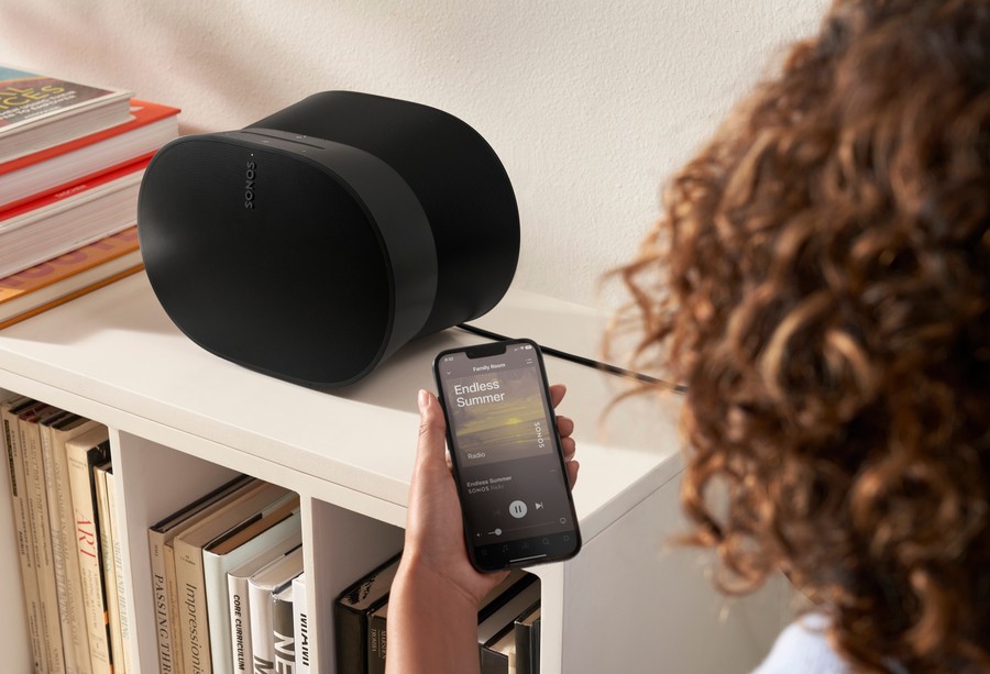 A woman uses an app on her smartphone to control the music on a black Sonos speaker situated on a white credenza.